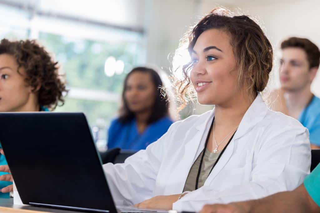 female medical student smiles confidently while using a laptop in class. Diverse students are in the background