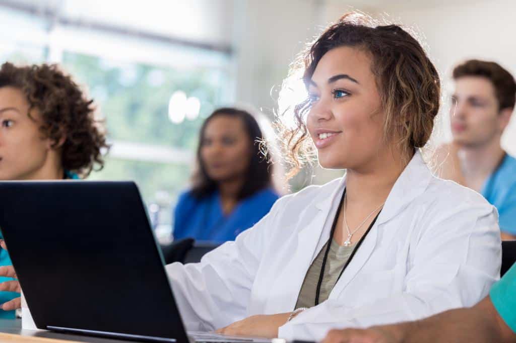 med student learning about what to expect when applying for a post-bacc program female medical student smiles confidently while using a laptop in class. Diverse students are in the background