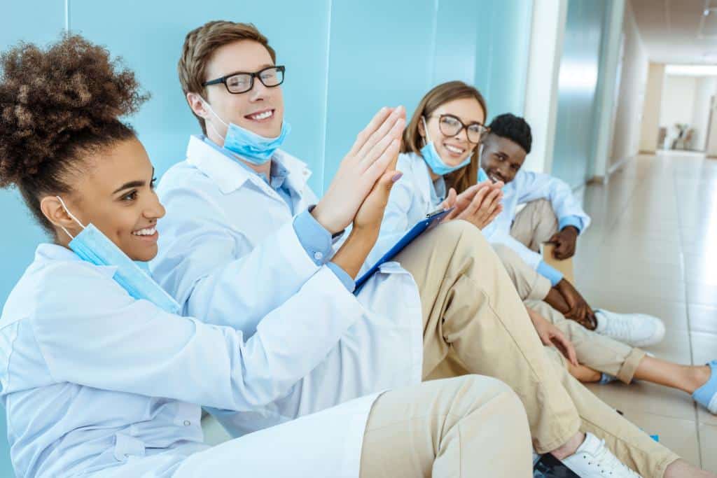 Smiling medical interns hanging out together on a floor in hospital four smiling young medical interns in white robes sitting on a floor in hospital
