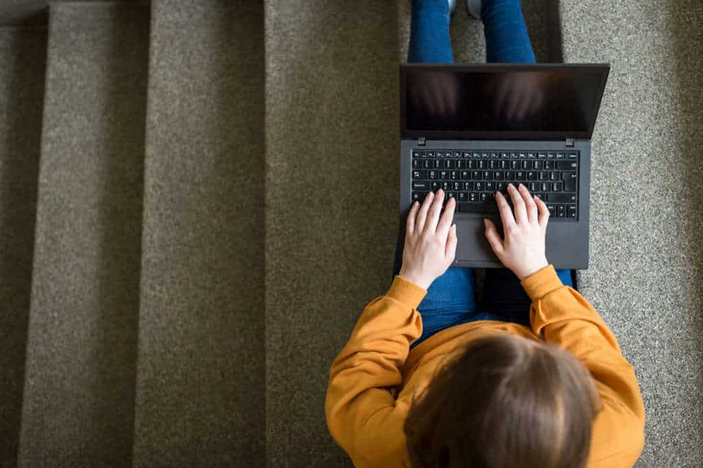 female college student sitting on stairs at school, writing essay on her laptop. Education concept.