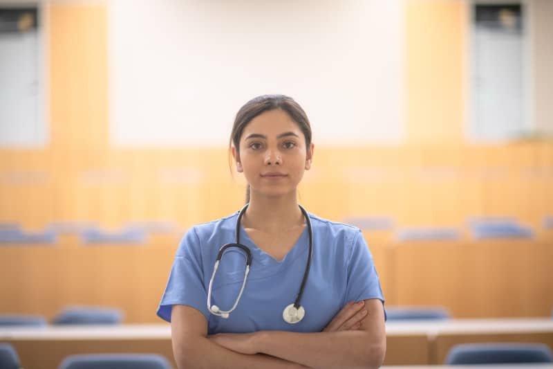 female Asian medical student poses in a lecture hall for a portrait wearing blue scrubs and stethoscope around her neck