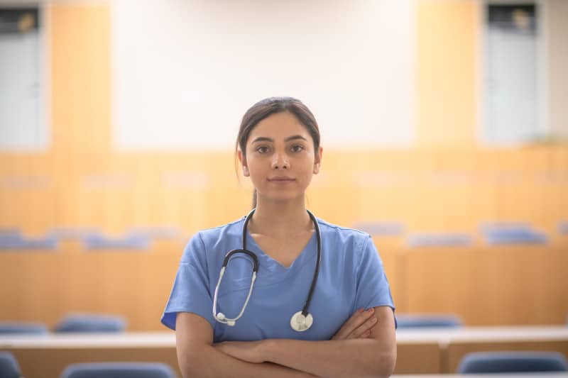 female medical student conveys need to research pre-medical post-bacc programs female Asian medical student poses in a lecture hall for a portrait wearing blue scrubs and stethoscope around her neck