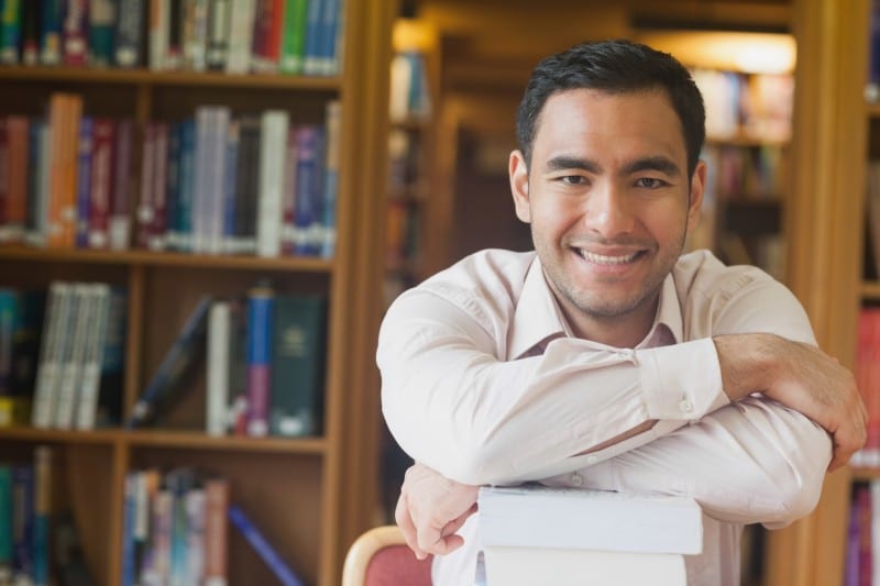 cheerful attractive man posing leaning on a stack of books in library smiling at camera